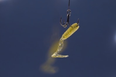 A close-up view of a fishing hook with a yellow, translucent artificial bait against a dark blue background. The bait has a curved tail and is slightly blurred, creating a sense of motion.