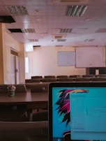 A lecture hall with multiple rows of empty seats and a projector screen at the front. The ceiling has several recessed light fixtures, and some panels appear to be missing. In the foreground, a laptop screen is visible displaying a colorful abstract wallpaper and a file explorer window with various document names.