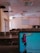 A lecture hall with multiple rows of empty seats and a projector screen at the front. The ceiling has several recessed light fixtures, and some panels appear to be missing. In the foreground, a laptop screen is visible displaying a colorful abstract wallpaper and a file explorer window with various document names.