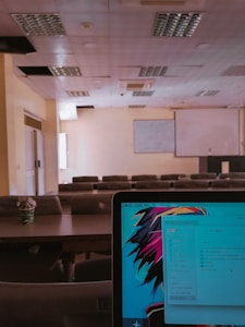 A lecture hall with multiple rows of empty seats and a projector screen at the front. The ceiling has several recessed light fixtures, and some panels appear to be missing. In the foreground, a laptop screen is visible displaying a colorful abstract wallpaper and a file explorer window with various document names.