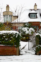 A house with a snow-covered roof and surrounding garden. The garden has a brick wall and a wrought iron gate, with snow also covering the ground and plants. Bare tree branches and evergreen shrubs are visible amidst the snow.