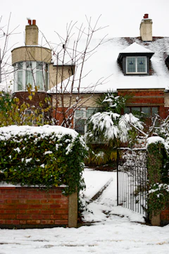 A house with a snow-covered roof and surrounding garden. The garden has a brick wall and a wrought iron gate, with snow also covering the ground and plants. Bare tree branches and evergreen shrubs are visible amidst the snow.