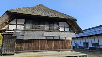 A traditional building with a thatched roof and wooden exterior. The structure features a balcony with wooden railings and opaque sliding windows. Adjacent to it, there's another building with a more modern metallic roof.