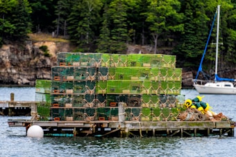 A stack of lobster traps is arranged on a wooden dock by the water. The traps are primarily green with some orange highlights, and there are ropes and buoys next to them. A small sailboat is anchored nearby, with a rocky, tree-lined shoreline in the background.