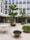An urban courtyard featuring modern architecture with large windows and a clean, minimalist design. The space includes several planter pots with green plants and trees, as well as a white metal table and chairs. The surroundings indicate a calm and organized atmosphere, likely part of an office or commercial building.