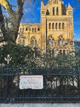 A historic building with ornate architectural features stands behind a wrought iron fence. The stone facade is highlighted by sunlight, showcasing intricate details and arches. A street sign reads 'The Royal Borough of Kensington and Chelsea, Cromwell Road, S.W.7', surrounded by leafy branches from nearby trees.