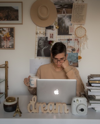 A person is seated at a desk with a laptop, surrounded by personal items and decorations. The desk holds a mug with a decorative moon, a small Eiffel Tower replica, a 'dream' sign, and an instant camera. The wall behind features a collage of photographs, a straw hat, and a dreamcatcher, creating a cozy and creative atmosphere.