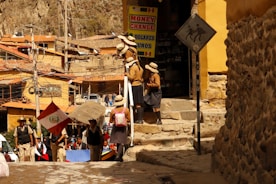 A group of people are standing near a building with a 'Money Change' sign. They appear to be wearing school uniforms with hats. A Peruvian flag is visible among them. The environment is urban and rustic with stone walls and buildings constructed with clay or similar materials. Electrical wires and poles are present, and there is a sign showing a person with a pack animal, suggesting a historical or tourist location.