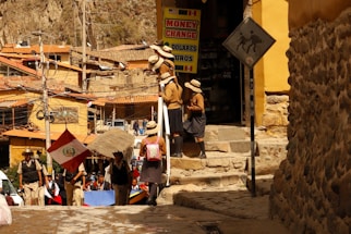 A group of people are standing near a building with a 'Money Change' sign. They appear to be wearing school uniforms with hats. A Peruvian flag is visible among them. The environment is urban and rustic with stone walls and buildings constructed with clay or similar materials. Electrical wires and poles are present, and there is a sign showing a person with a pack animal, suggesting a historical or tourist location.