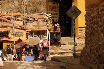 A group of people are standing near a building with a 'Money Change' sign. They appear to be wearing school uniforms with hats. A Peruvian flag is visible among them. The environment is urban and rustic with stone walls and buildings constructed with clay or similar materials. Electrical wires and poles are present, and there is a sign showing a person with a pack animal, suggesting a historical or tourist location.