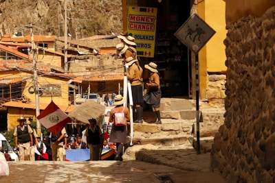A group of people are standing near a building with a 'Money Change' sign. They appear to be wearing school uniforms with hats. A Peruvian flag is visible among them. The environment is urban and rustic with stone walls and buildings constructed with clay or similar materials. Electrical wires and poles are present, and there is a sign showing a person with a pack animal, suggesting a historical or tourist location.