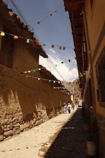 A narrow, cobblestone alley bordered by adobe walls and lined with colorful flags and decorations overhead. The clear blue sky provides a bright backdrop, while several people walk leisurely along the path. The buildings have rustic, traditional architecture with wooden elements and clay roof tiles.
