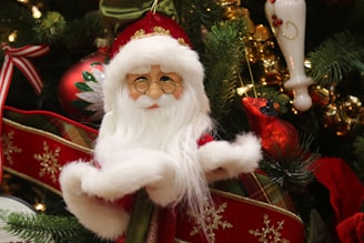 A Santa Claus ornament with a white beard and round glasses is placed among Christmas tree branches and decorated with a variety of festive ornaments, including a red cardinal, ribbons, and a hand-painted bauble.