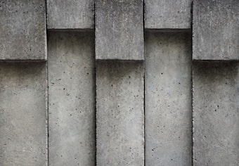 Concrete wall with vertical columns and a textured surface. The pattern is formed by repeating rectangular shapes in a symmetrical arrangement. Small pockmarks and varying shades of gray contribute to the industrial appearance.