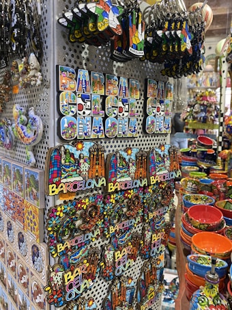 A display of colorful souvenirs featuring the word 'Barcelona' in various designs, alongside vibrant ceramic items. The image showcases keychains, magnets, and decorative pieces with artistic representations of landmarks. Rows of brightly painted bowls and dishes add to the vibrant atmosphere.