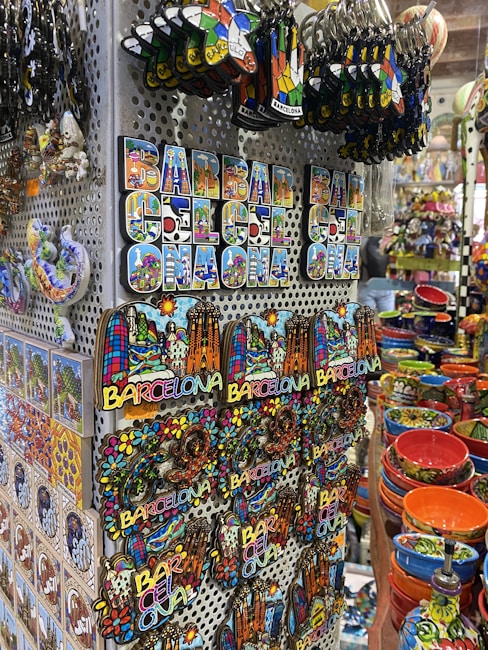 A display of colorful souvenirs featuring the word 'Barcelona' in various designs, alongside vibrant ceramic items. The image showcases keychains, magnets, and decorative pieces with artistic representations of landmarks. Rows of brightly painted bowls and dishes add to the vibrant atmosphere.