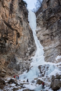 A towering frozen waterfall cascades down a steep, rocky cliff face. The rugged terrain is covered with patches of snow, and several climbers equipped with ice axes and helmets are seen making their way up the icy surface. The bare trees at the top hint at a cold, wintry environment.