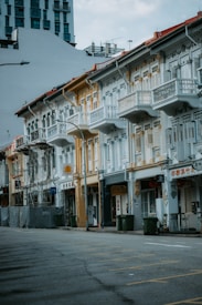 A row of charming, well-preserved shophouses with ornate facades and shuttered windows. The buildings feature a blend of pastel blue, white, and yellow colors. The street is empty, and a few trash bins are placed along the sidewalk. A building under construction is visible to the left, covered in scaffolding.