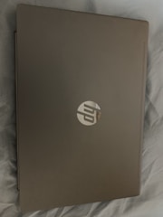 A closed laptop with a metallic finish, displaying a logo on the top. The laptop is placed on a soft, light-colored fabric surface.