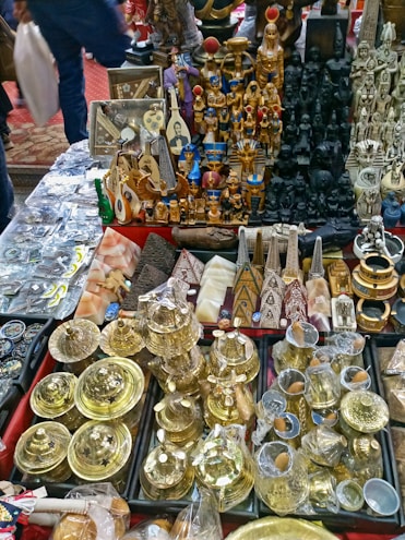 A collection of various souvenirs and artifacts, including golden pots, statuettes resembling ancient Egyptian figures, pyramids, and other intricately designed decorative items spread across a table. The assortment displays a mix of metal, wood, and stone objects with a focus on Egyptian-themed cultural motifs.