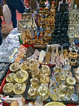 A collection of various souvenirs and artifacts, including golden pots, statuettes resembling ancient Egyptian figures, pyramids, and other intricately designed decorative items spread across a table. The assortment displays a mix of metal, wood, and stone objects with a focus on Egyptian-themed cultural motifs.