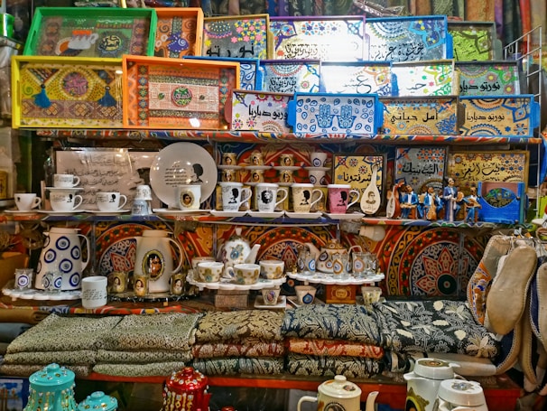 A vibrant collection of decorated trays, cups, teapots, and textiles is arranged on shelves. The trays feature intricate designs and patterns, some with Arabic script. There are also figures wearing traditional attire and various pottery pieces adorned with portraits and geometric motifs.