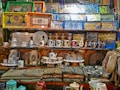 A vibrant collection of decorated trays, cups, teapots, and textiles is arranged on shelves. The trays feature intricate designs and patterns, some with Arabic script. There are also figures wearing traditional attire and various pottery pieces adorned with portraits and geometric motifs.