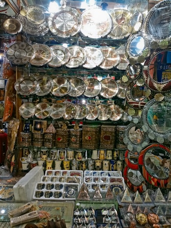 An artisanal display with intricately designed metal plates featuring elaborate engravings, ornate boxes, and various traditional items. The arrangement also includes small pyramidal souvenirs and mugs with decorative motifs, set against a glass shelf backdrop.