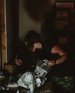 A person is working in a dimly lit workshop, handling parts of an engine. They are focused, with one hand holding a tool and the other stabilizing the machinery. The surroundings are cluttered with mechanical components and tools. Shelves filled with boxes and equipment are visible in the background.