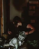 A person is working in a dimly lit workshop, handling parts of an engine. They are focused, with one hand holding a tool and the other stabilizing the machinery. The surroundings are cluttered with mechanical components and tools. Shelves filled with boxes and equipment are visible in the background.