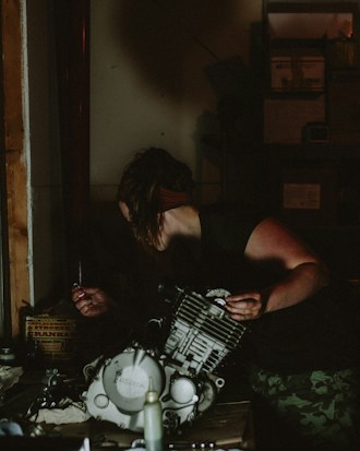 A person is working in a dimly lit workshop, handling parts of an engine. They are focused, with one hand holding a tool and the other stabilizing the machinery. The surroundings are cluttered with mechanical components and tools. Shelves filled with boxes and equipment are visible in the background.
