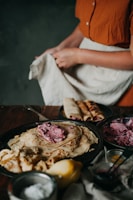A person is preparing a dish with several crepes on a black plate, topped with a dollop of pink creamy filling. Beside them are neatly rolled crepes, along with small bowls containing various ingredients. A lemon and a container with powdered sugar are also visible. The person is holding a cloth and wearing a rust-colored shirt.