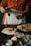 A person in an orange shirt and white apron is pouring dark red syrup onto a stack of crepes on a wooden table. There are additional crepes on a plate in the background and various kitchen items, including a lemon and a small container of salt.