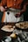 A person in an orange shirt and white apron is pouring dark red syrup onto a stack of crepes on a wooden table. There are additional crepes on a plate in the background and various kitchen items, including a lemon and a small container of salt.