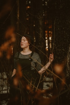 A person with closed eyes stands peacefully in a forest, leaning against a tree. They are wearing a dark outfit, and the atmosphere is serene and introspective. The background is composed of tall trees with dark bark and sparse branches.
