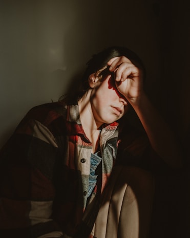 A person sitting in a dimly lit space with one hand covering their face. The person is wearing a plaid shirt and appears to be in a contemplative or introspective mood. Light and shadow play across their face, emphasizing the sense of solitude.