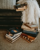 A cozy corner with a stack of various books placed on the carpeted floor, next to a plush cushion and a fluffy white throw over a dark leather couch. The books range from modern hardcovers to worn vintage editions, creating a warm, inviting ambiance.