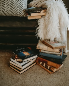 A cozy corner with a stack of various books placed on the carpeted floor, next to a plush cushion and a fluffy white throw over a dark leather couch. The books range from modern hardcovers to worn vintage editions, creating a warm, inviting ambiance.