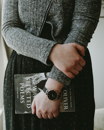 A person is holding a book titled 'New and Selected Poems' while wearing a dark-colored watch. The person is dressed in a grey textured sweater and a dark skirt with subtle patterns. The framing suggests a calm and introspective moment.