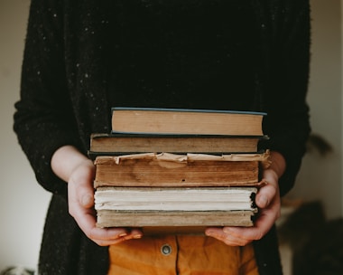 A person holding a stack of old, worn books with visible tears and discoloration on the pages and covers. The hands are partially visible, wearing casual clothing, with a dark sweater. The background is softly blurred, placing emphasis on the books.