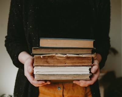 A person holding a stack of old, worn books with visible tears and discoloration on the pages and covers. The hands are partially visible, wearing casual clothing, with a dark sweater. The background is softly blurred, placing emphasis on the books.