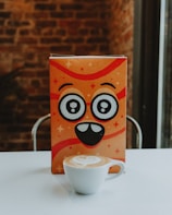 A playful, cartoonish orange box with large, whimsical eyes and a smiling mouth is centered on a white surface. In front of the box is a white cup of cappuccino with latte art on top. The background consists of a blurred brick wall, suggesting a cozy indoor setting.