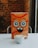 A playful, cartoonish orange box with large, whimsical eyes and a smiling mouth is centered on a white surface. In front of the box is a white cup of cappuccino with latte art on top. The background consists of a blurred brick wall, suggesting a cozy indoor setting.