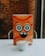 A playful, cartoonish orange box with large, whimsical eyes and a smiling mouth is centered on a white surface. In front of the box is a white cup of cappuccino with latte art on top. The background consists of a blurred brick wall, suggesting a cozy indoor setting.