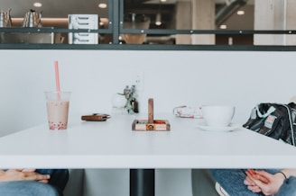 A white table in a cafe setting, featuring a bubble tea with a red straw, a white coffee cup on a saucer, a wooden puzzle, and a black bag. A few other items, like a wallet and a small vase, are also present. The background includes shelves with stainless steel containers.