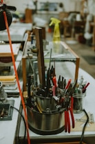 A workbench cluttered with various hand tools, including pliers, scissors, and files, organized in a cylindrical holder. A yellow spray bottle sits in the background alongside other miscellaneous items, suggesting a workshop environment.