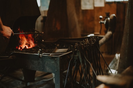 A blacksmith's forge with tools hanging in the foreground, glowing coals in a hearth, and a person's hand tending to the fire. The setting appears rustic and workshop-like, with a focus on the traditional craft of metalworking.