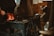 A blacksmith's forge with tools hanging in the foreground, glowing coals in a hearth, and a person's hand tending to the fire. The setting appears rustic and workshop-like, with a focus on the traditional craft of metalworking.