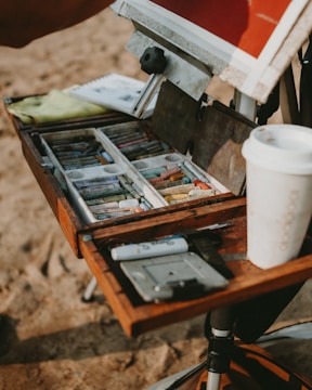 A close-up of an artist's easel and supply box set up on a sandy surface. The wooden box contains various colored pastels neatly arranged in compartments. A sketchpad and a white takeaway coffee cup are also visible. The scene suggests an outdoor painting session.