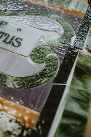 A close-up of a stained glass artwork with intricate patterns and text. The image features a mix of smooth and textured glass pieces in various colors, arranged in a detailed design.